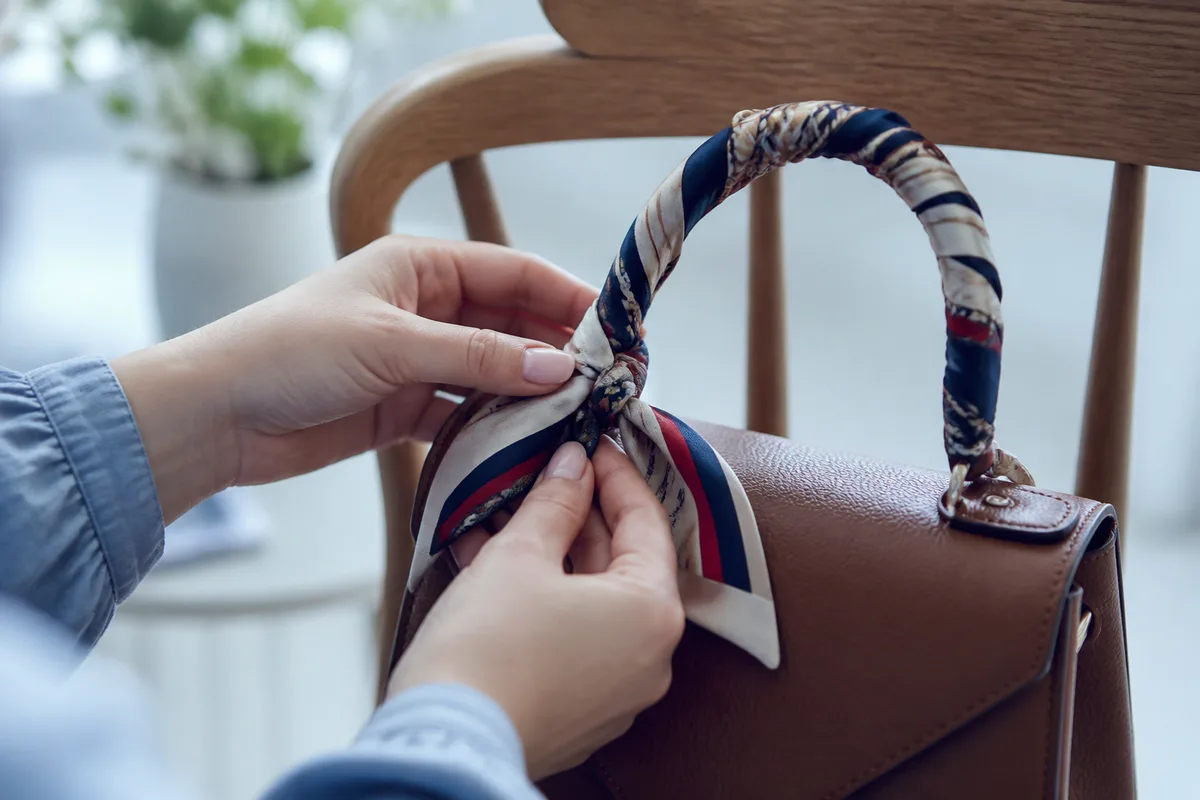 Carré de soie noué sur l’anse d’un sac en cuir posé sur une chaise